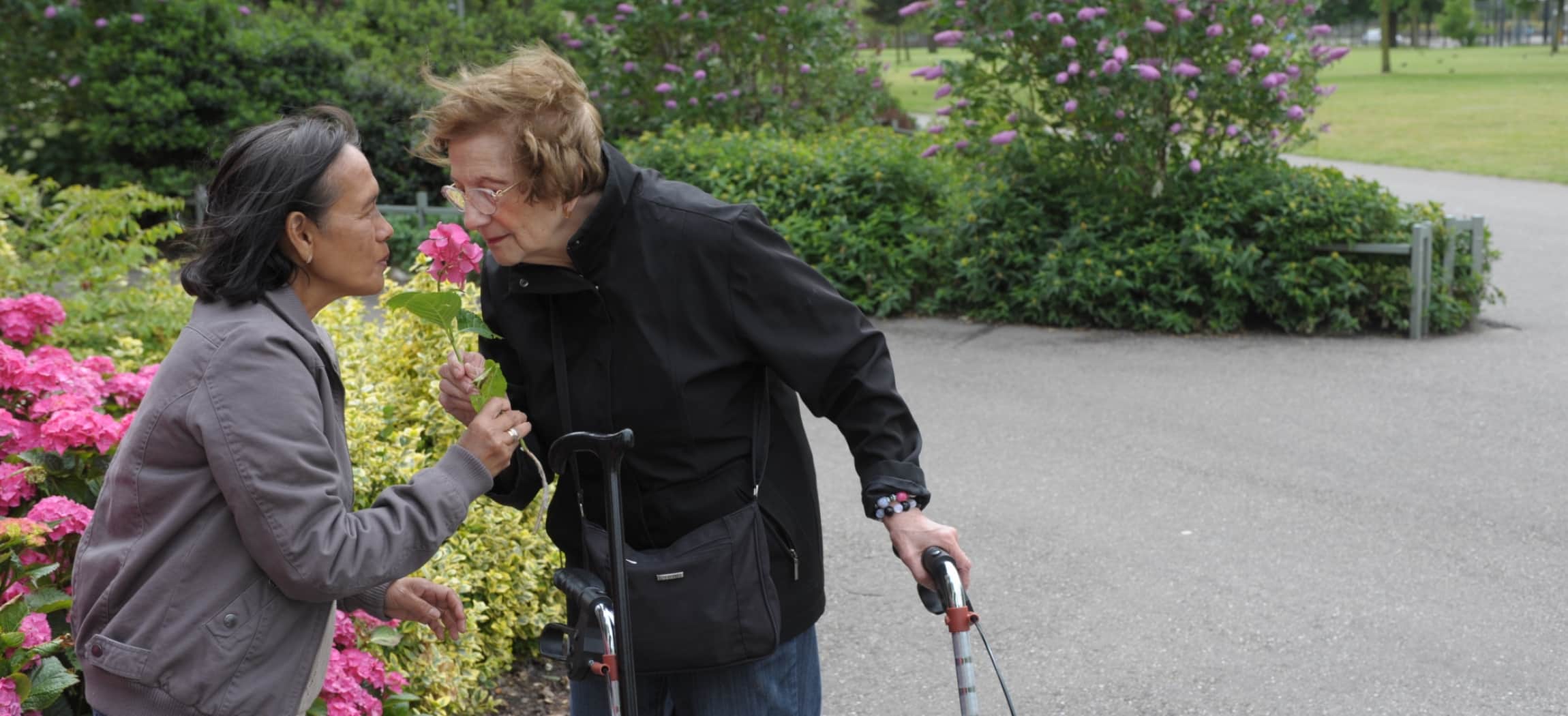Vrouw met rollator buiten in een park ruikt aan een bloem samen met andere vrouw