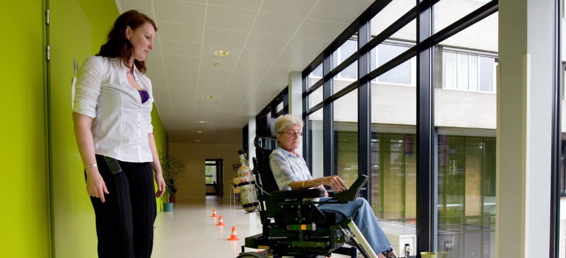 Vrouw in elektrische rolstoel oefent in lange gang met pylon pilon met ergotherapeut