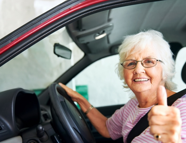 Vrouw grijs haar met bril zit achter het stuur in een auto met duim omhoog